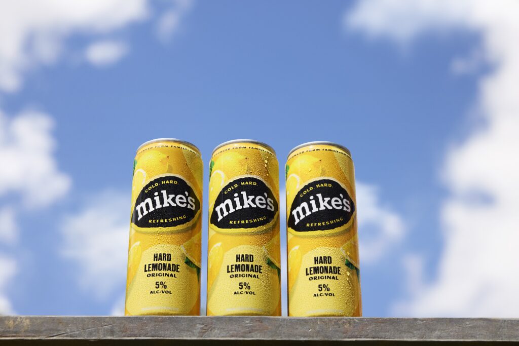 Three chilled, condensation-covered cans of Mike’s Hard Lemonade standing side-by-side against a vibrant blue sky with scattered white clouds. The yellow cans feature the 'mike's' logo on a black lemon-shaped background, with 'COLD • HARD • REFRESHING' and 'HARD LEMONADE' visible.